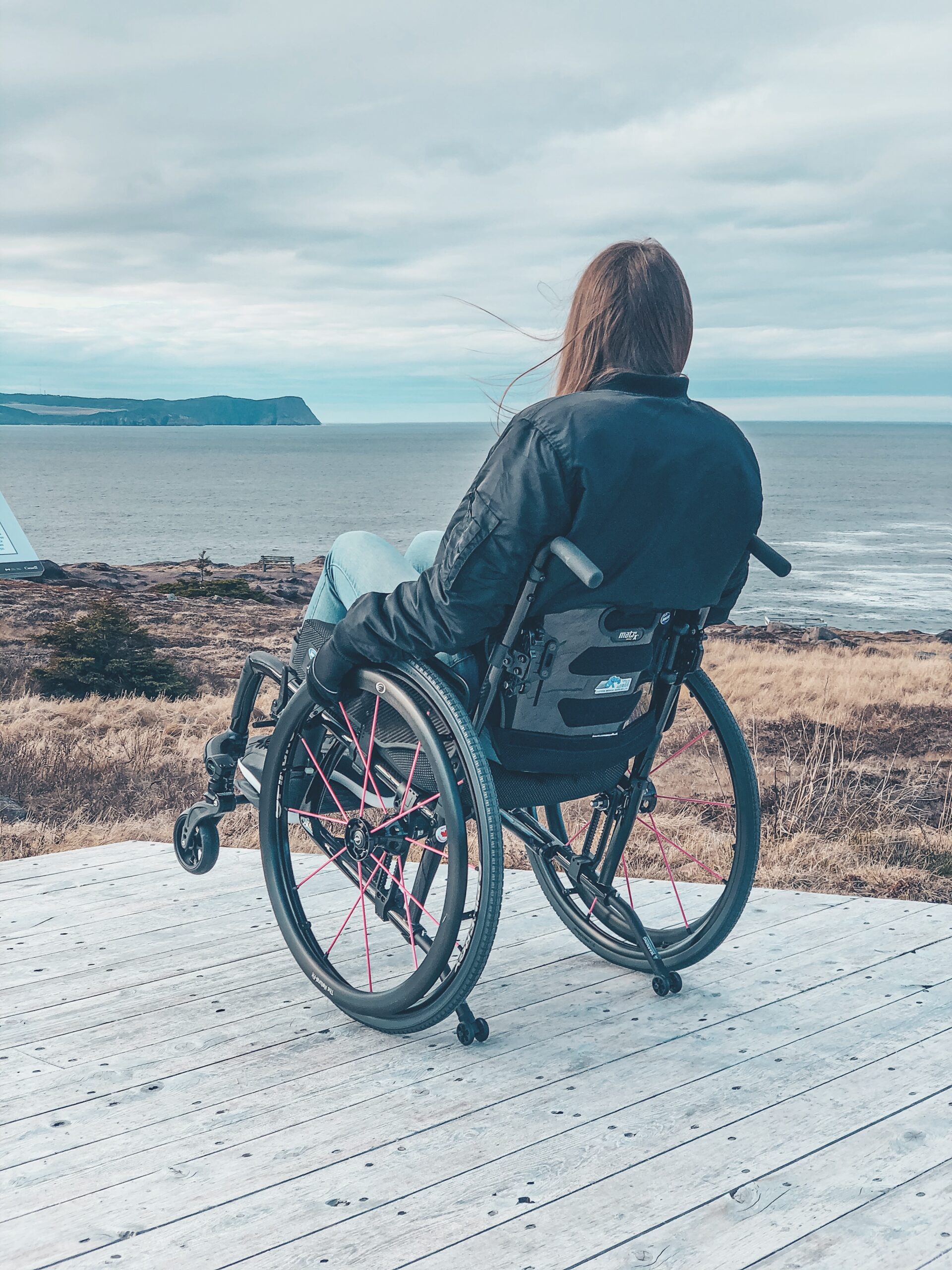 A photo of me from the back, sitting in my wheelchair looking out at the rocks and ocean from a wooden observation deck at Cape Spear. Photo credit: Matt McCoag
