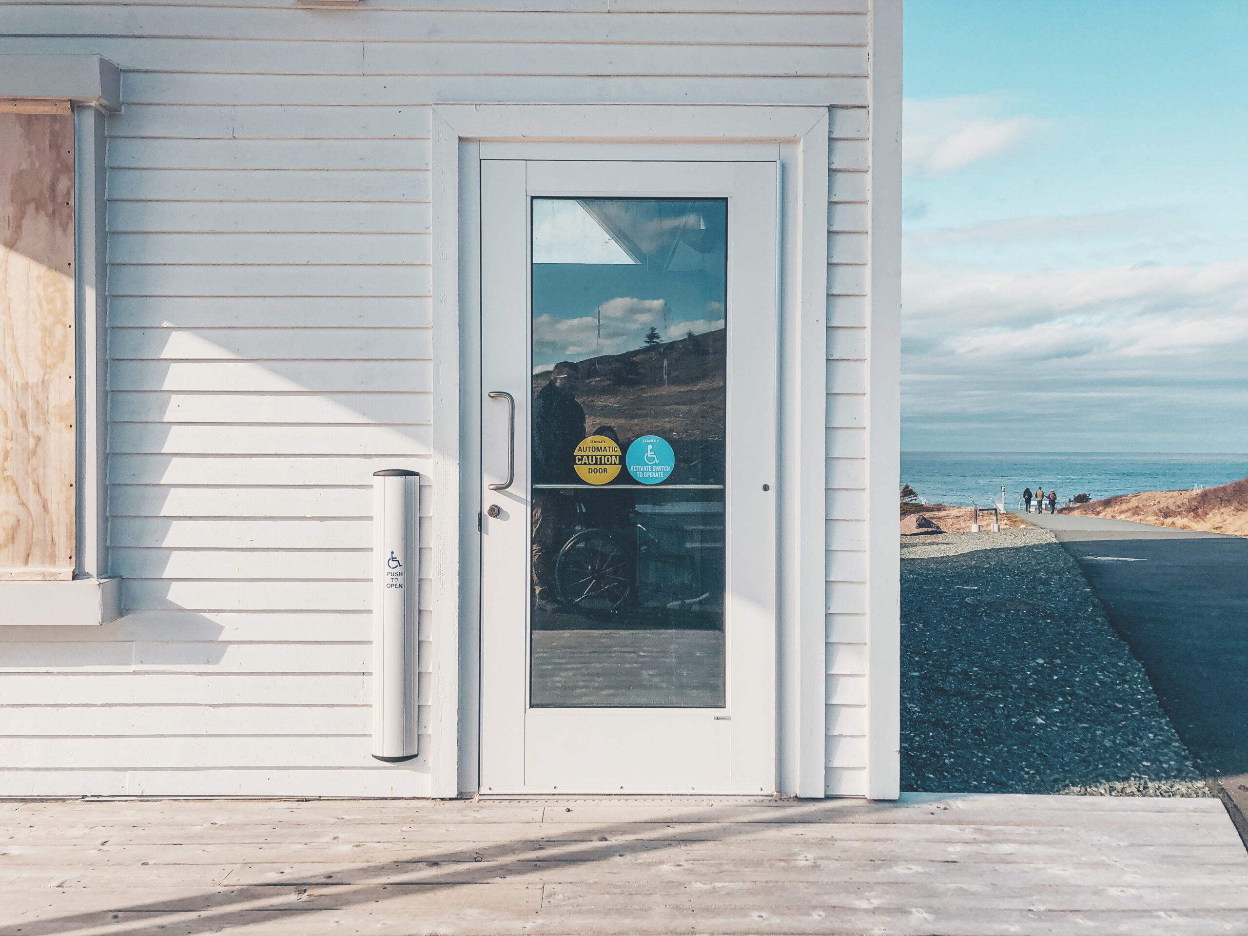 A photo of a white building with a ramp leading up to it. It houses the public washrooms and on one door there is an accessibility sign.