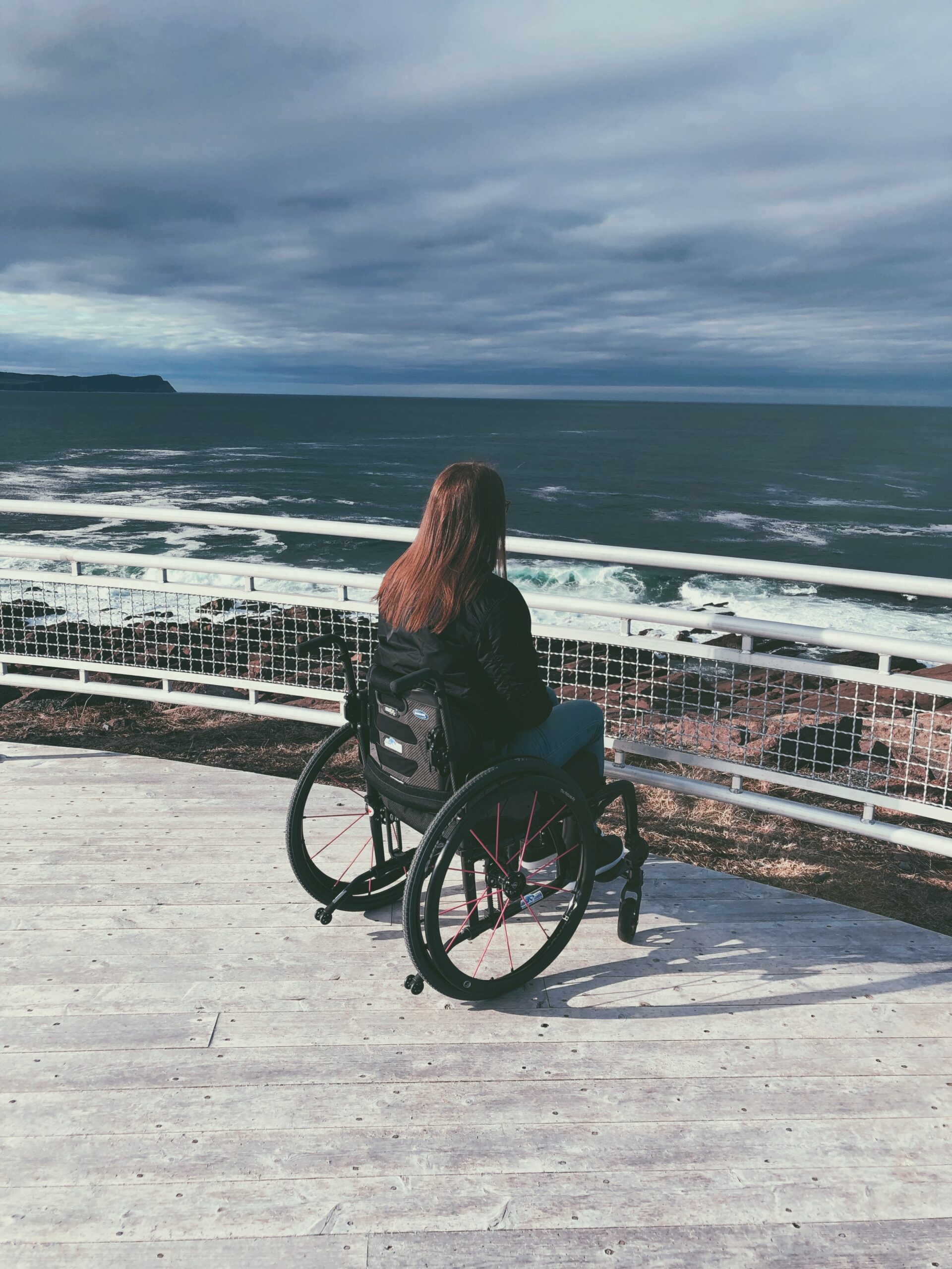 A photo of me from behind, sitting in my wheelchair on a different observation deck near the end of the trail. There are rails in front of me and then cliffs and ocean beyond.