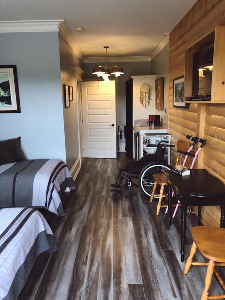 Interior view of the accessible suite. The right wall is wooden and has two chairs, a small black table, kitchenette and closet. The left side is painted blue and you can see the ends of two beds as well as the bathroom door beyond that.