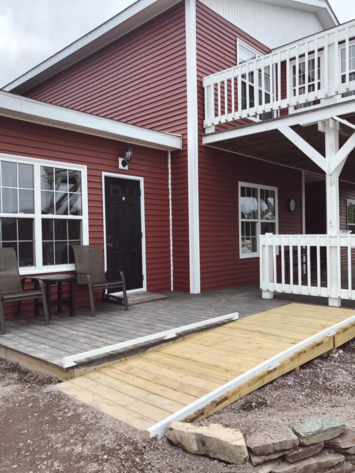 A photo of the exterior of the accessible suite, with a ramp leading to a deck with two chairs and a table in front of a large window, and a black door with a small ramp.