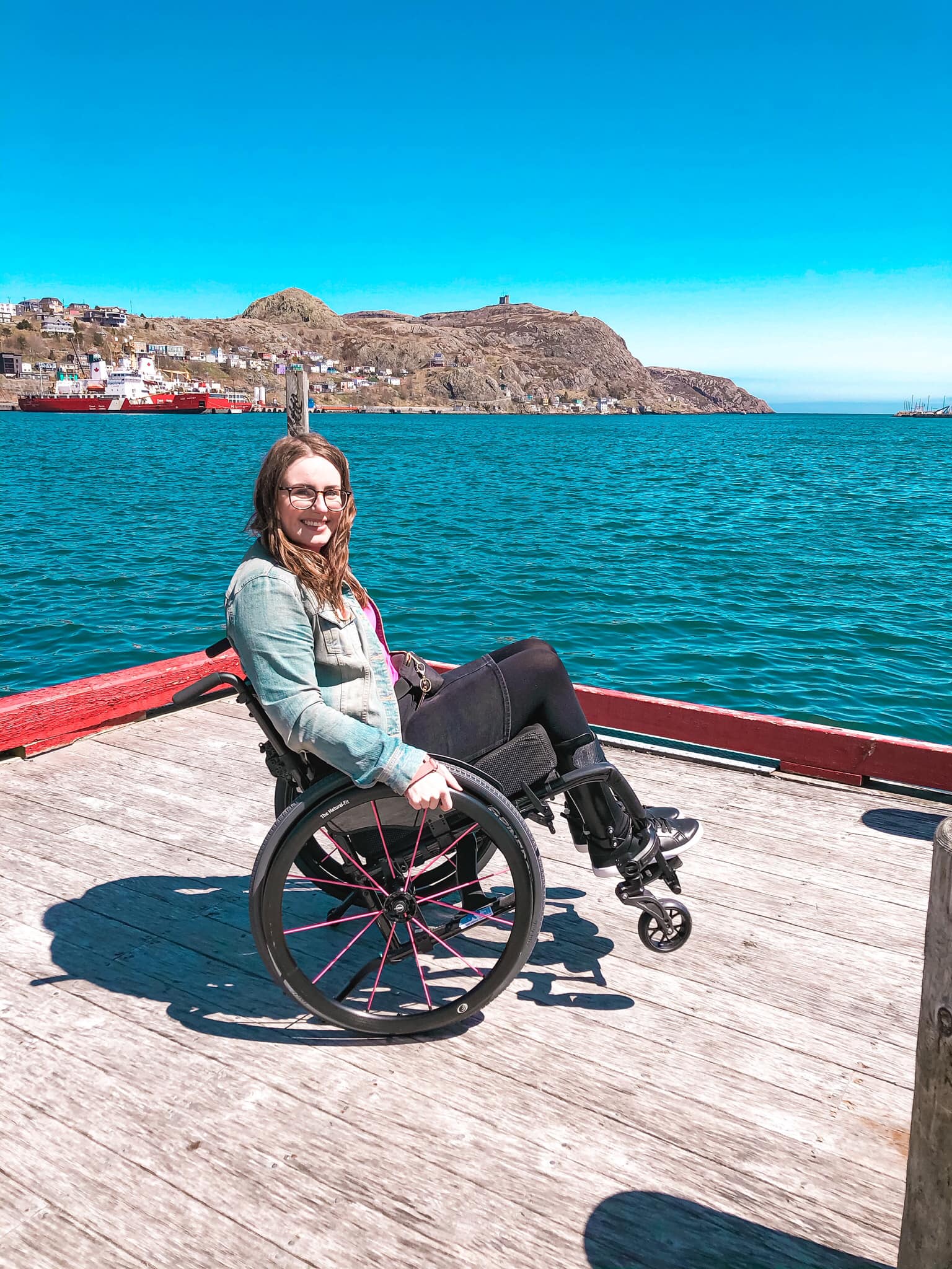 A photo of me doing a wheelie in my wheelchair on the wharf at Harbourside Park. Behind me is the St. John’s Harbour, Signal Hill and the Battery in the distance, and blue sky above.
