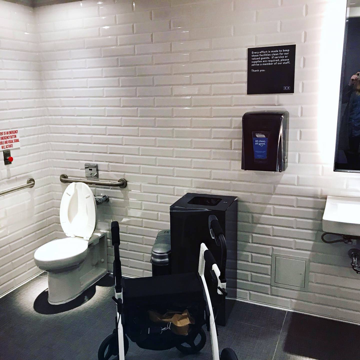 The interior of the accessible washroom which shows a large white room with railing, a toilet, and a sink that you can wheel under.
