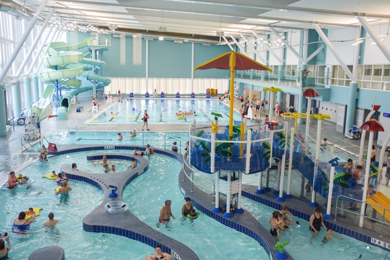 A photo of the pool area at Paul Reynolds Community Centre. In the foreground is the lazy river and children's splash pad, and in the background is the larger pool and water slides.