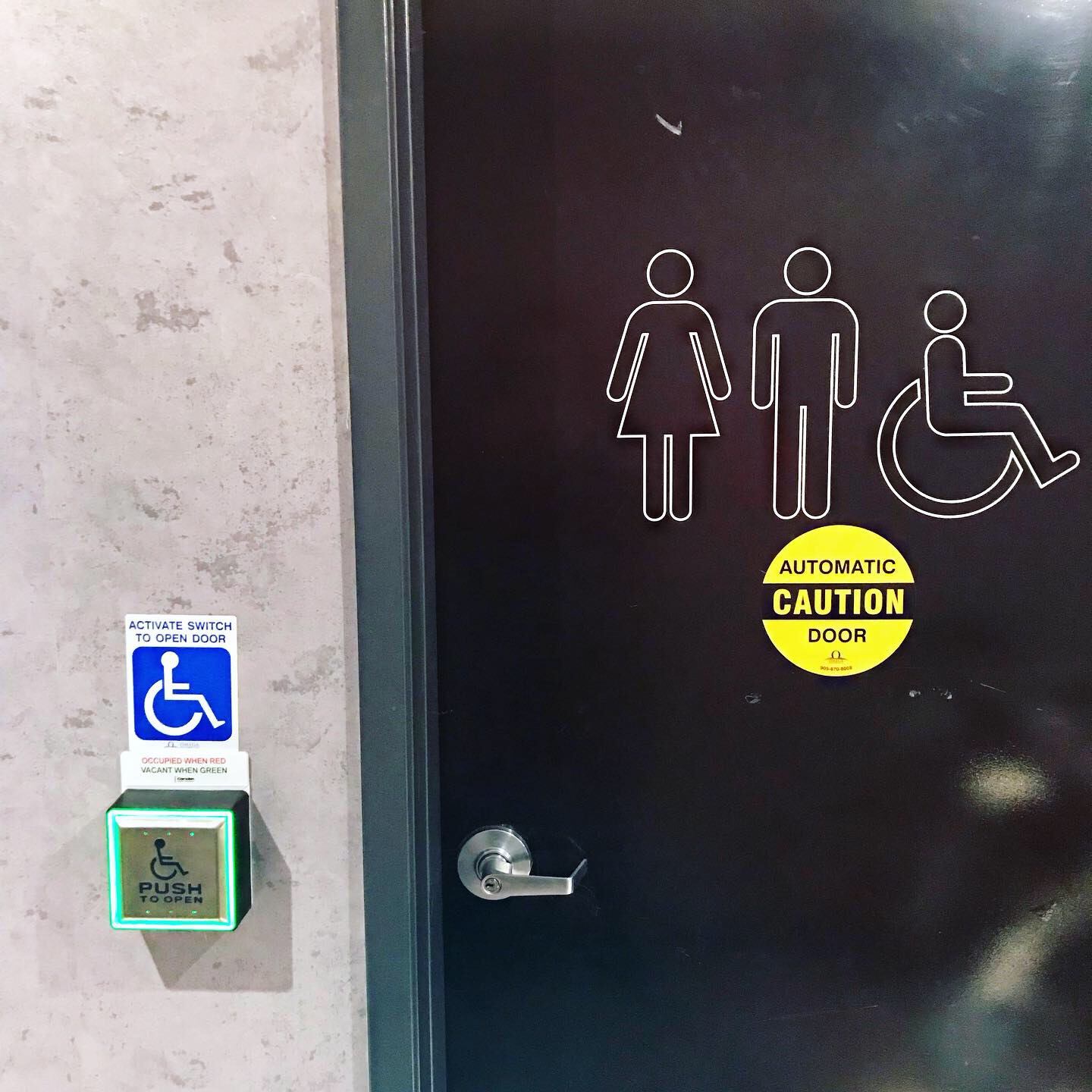 An accessible washroom door. On the left is an automatic door button that is lit green because the washroom is vacant.