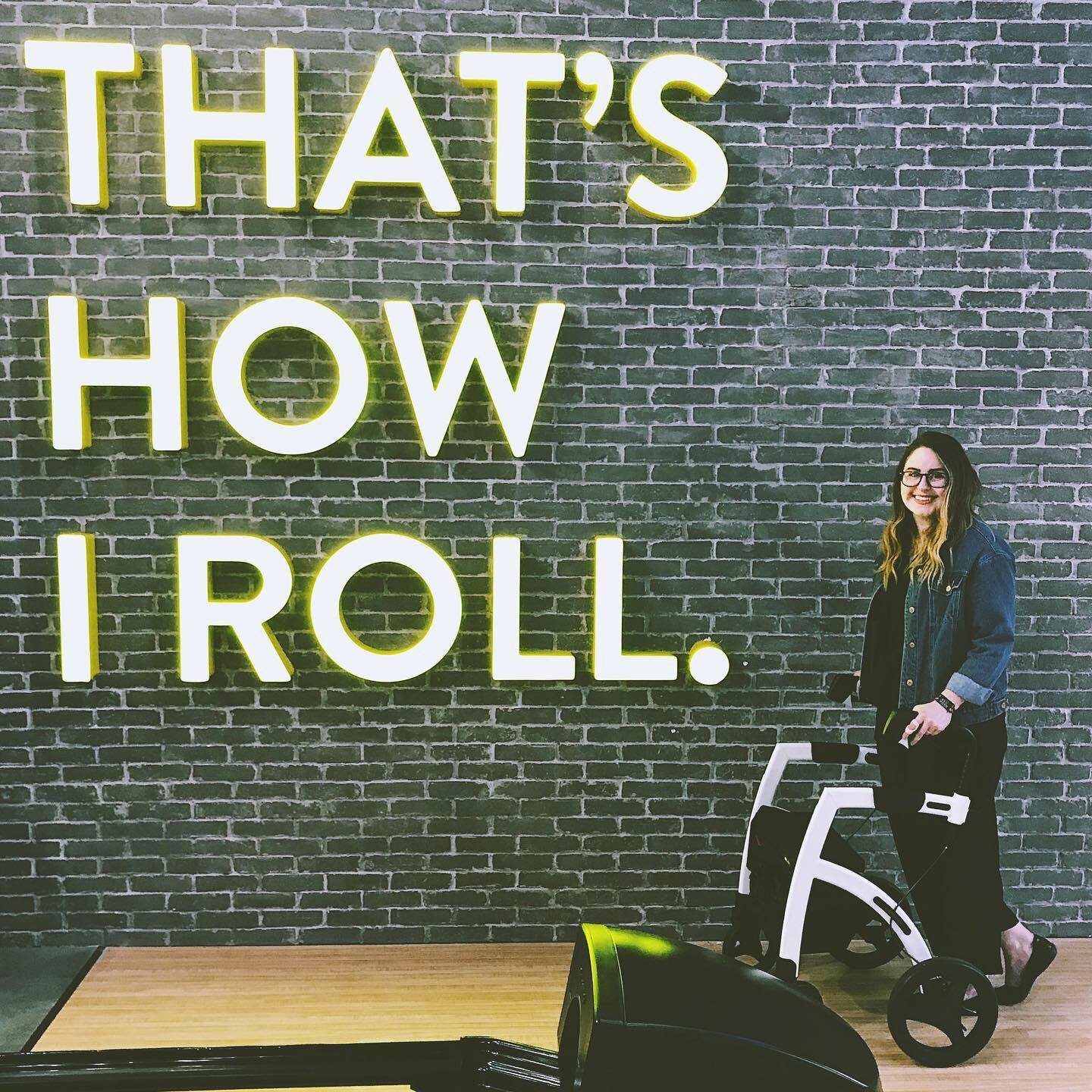 Lisa is standing with her white rollator in front of a brick wall with a neon sign that reads: That's How I Roll.