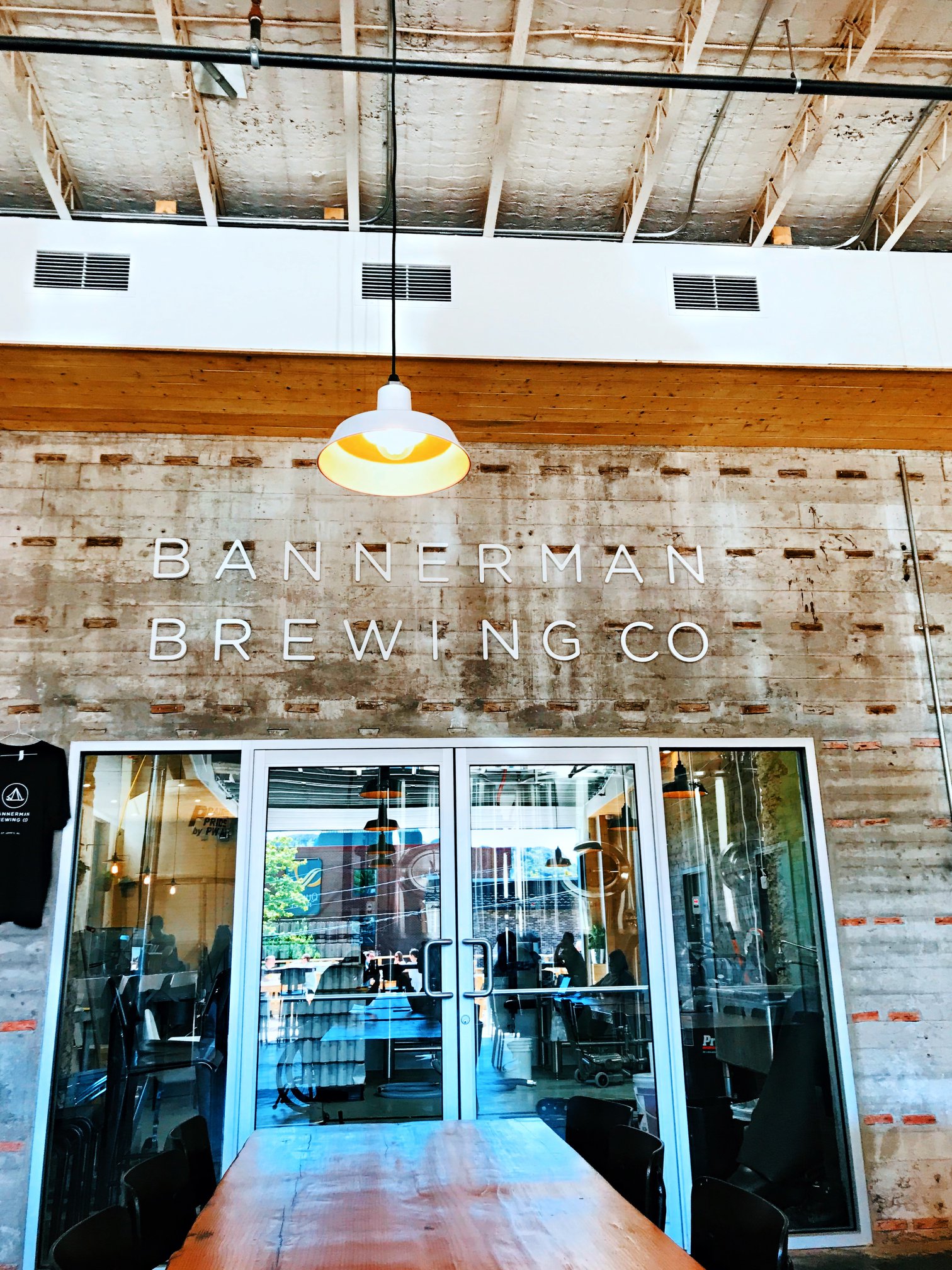 A photo of the back wall of Bannerman Brewing Co. At the top of the wood wall is a sign with the business name. Underneath the sign are glass doors, and through the glass there is brewing equipment visible.