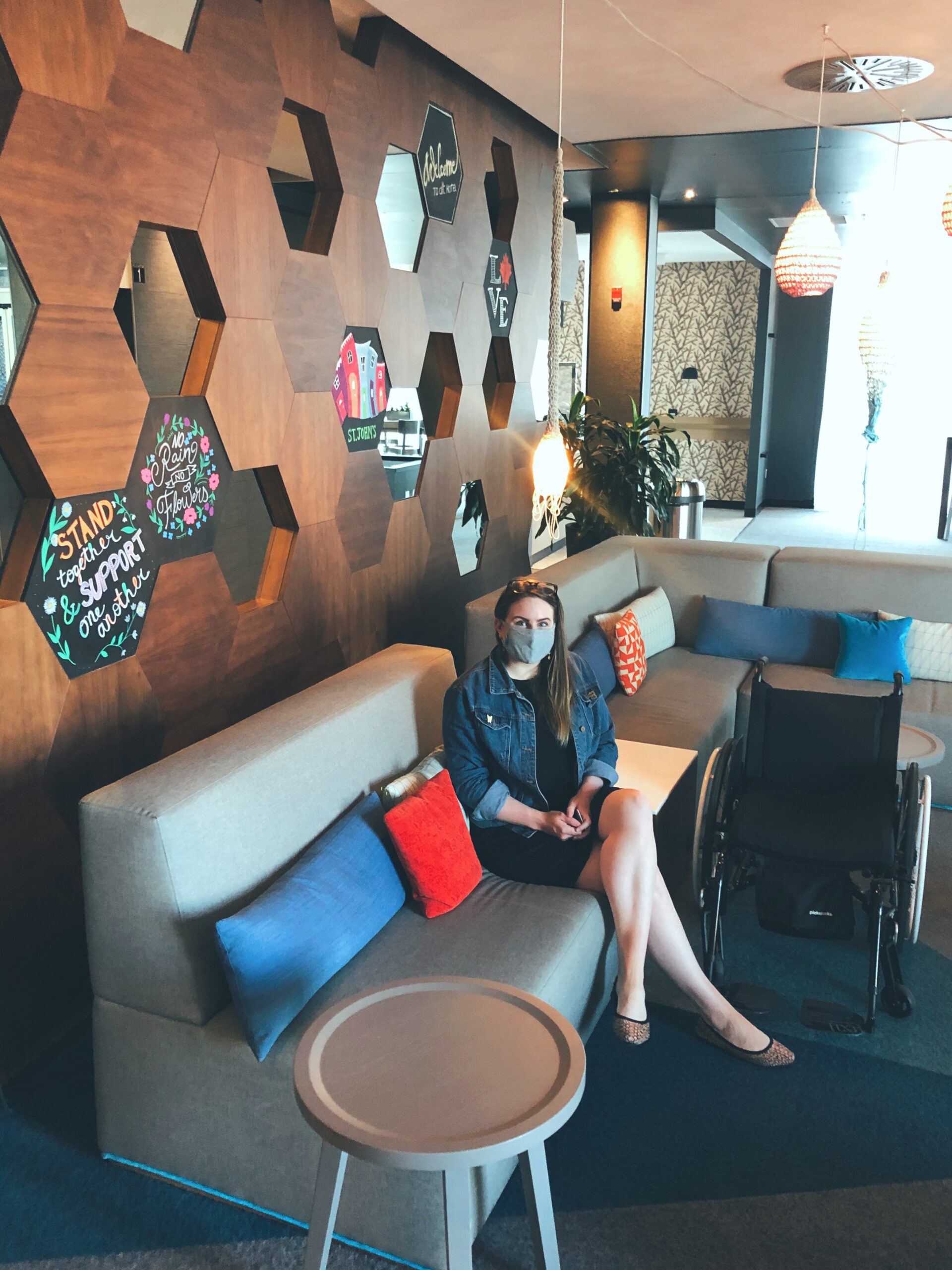 A photo of me in the Alt Hotel lobby. I'm sitting on a light grey sectional with blue, red and orange cushions. Next to the couch is my wheelchair and behind me is a very cool hexagonal wooden tiled wall. 