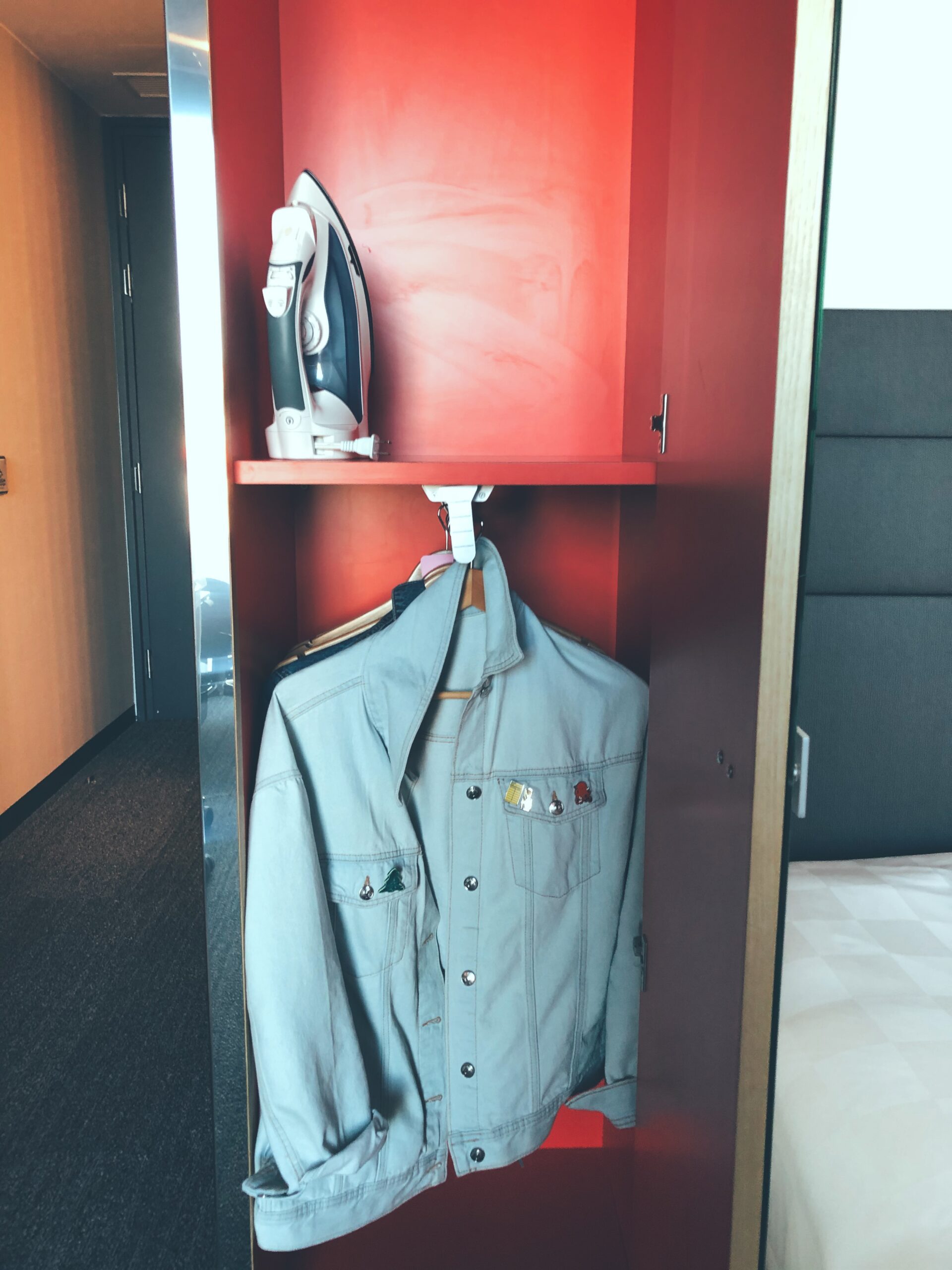 A photo of the inside of the closet in the accessible room. It is red and there is a road with a denim jacket hanging at wheelchair height and a shelf above with an iron.