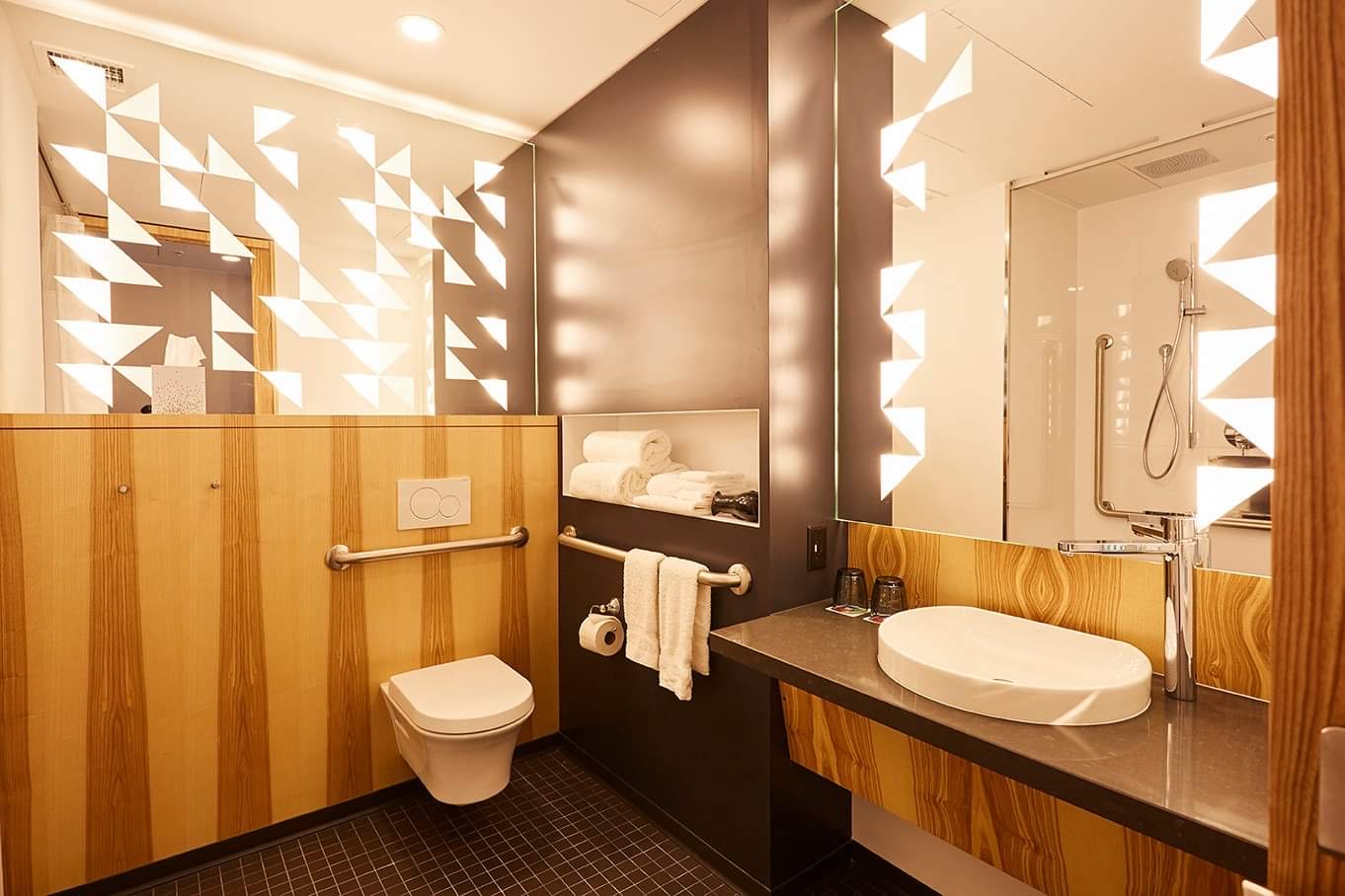 A photo of one side of the washroom in the accessible rooms at the Alt. Lots of space, a counter you can wheel under with a large mirror above it, a toilet in the corner with grab bars behind and to the side.