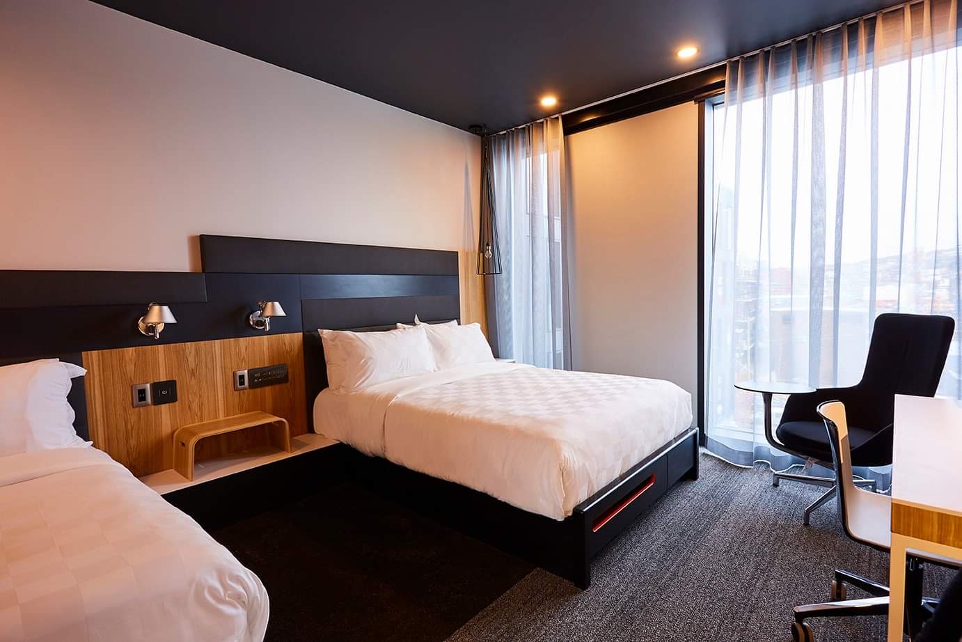 A photo of the interior of an accessible room at the Alt Hotel. A large window on the far wall, a desk and chair, a queen bed and a single bed with a nightstand in between the two.