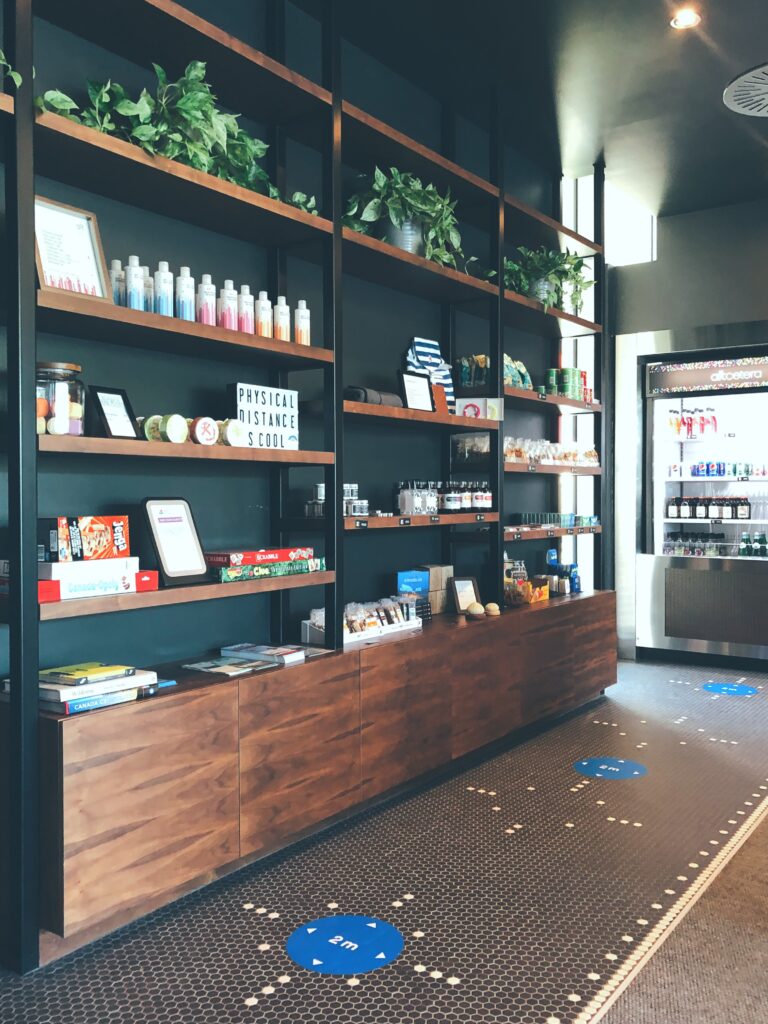 A photo of a wooden display shelf in the lobby of the Alt Hotel. On the shelves are some books, board games, and various merchandise for sale. In the background is a cooler with drinks.