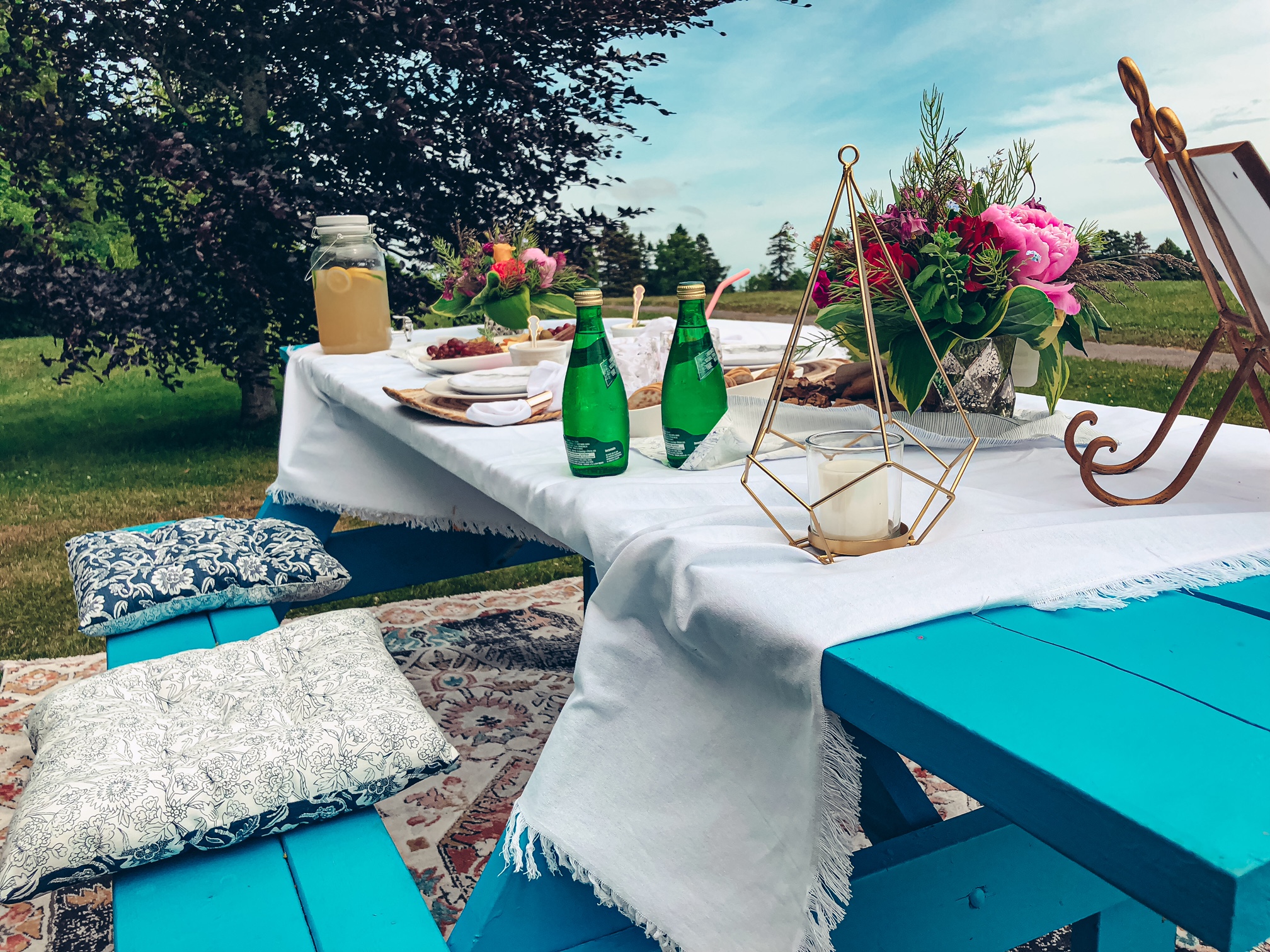 A close up photo of a bright blue picnic table with two cushions on the bench. On top of the table is a white linen tablecloth, two bouquets of flowers, several plates of food and drinks, and fancy dinnerware. 