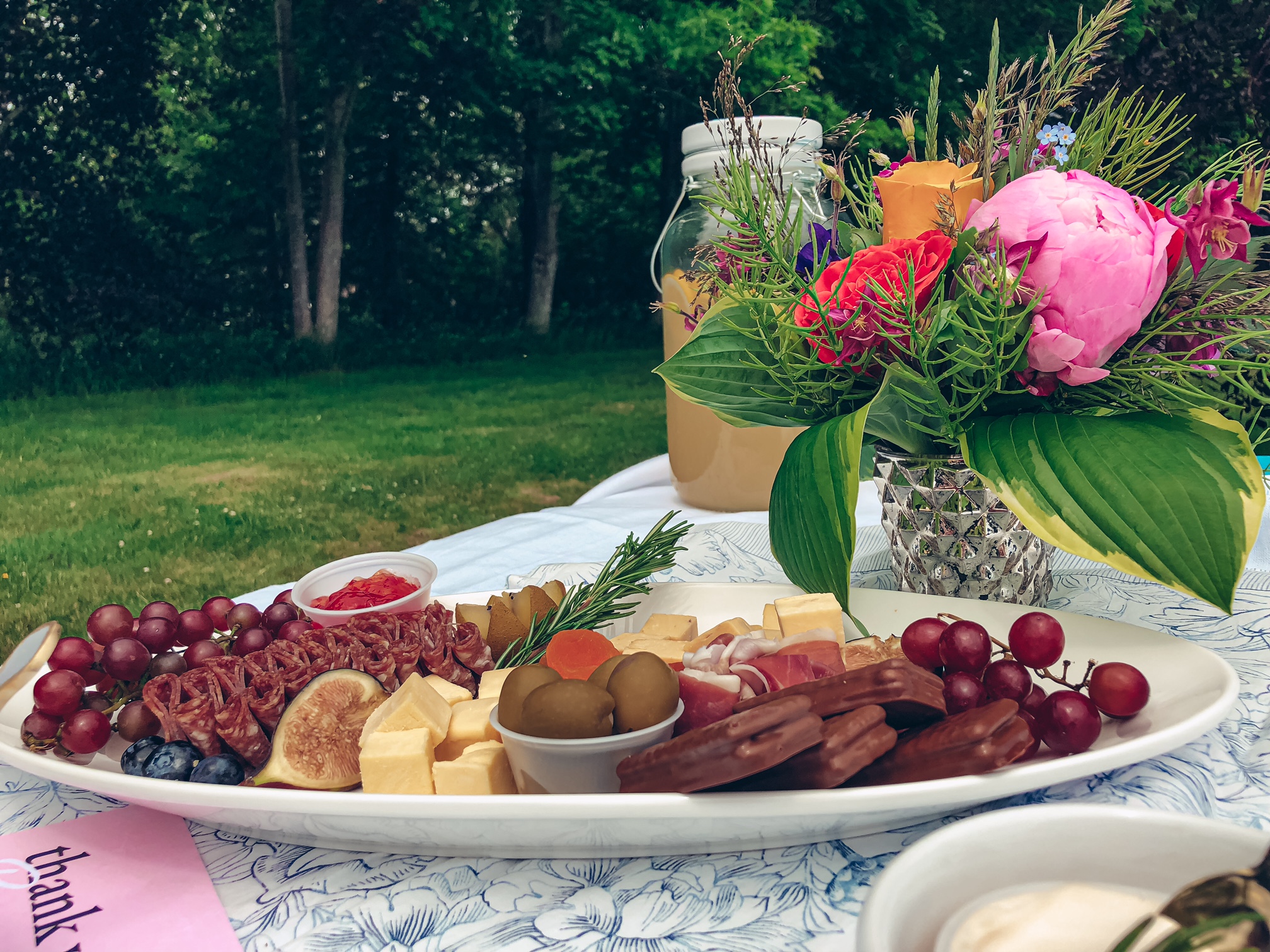 A close up of a white platter filled with charcuterie, resting on a white and blue table runner. Behind it is a bouquet of flowers and a pitcher of lemonade.
