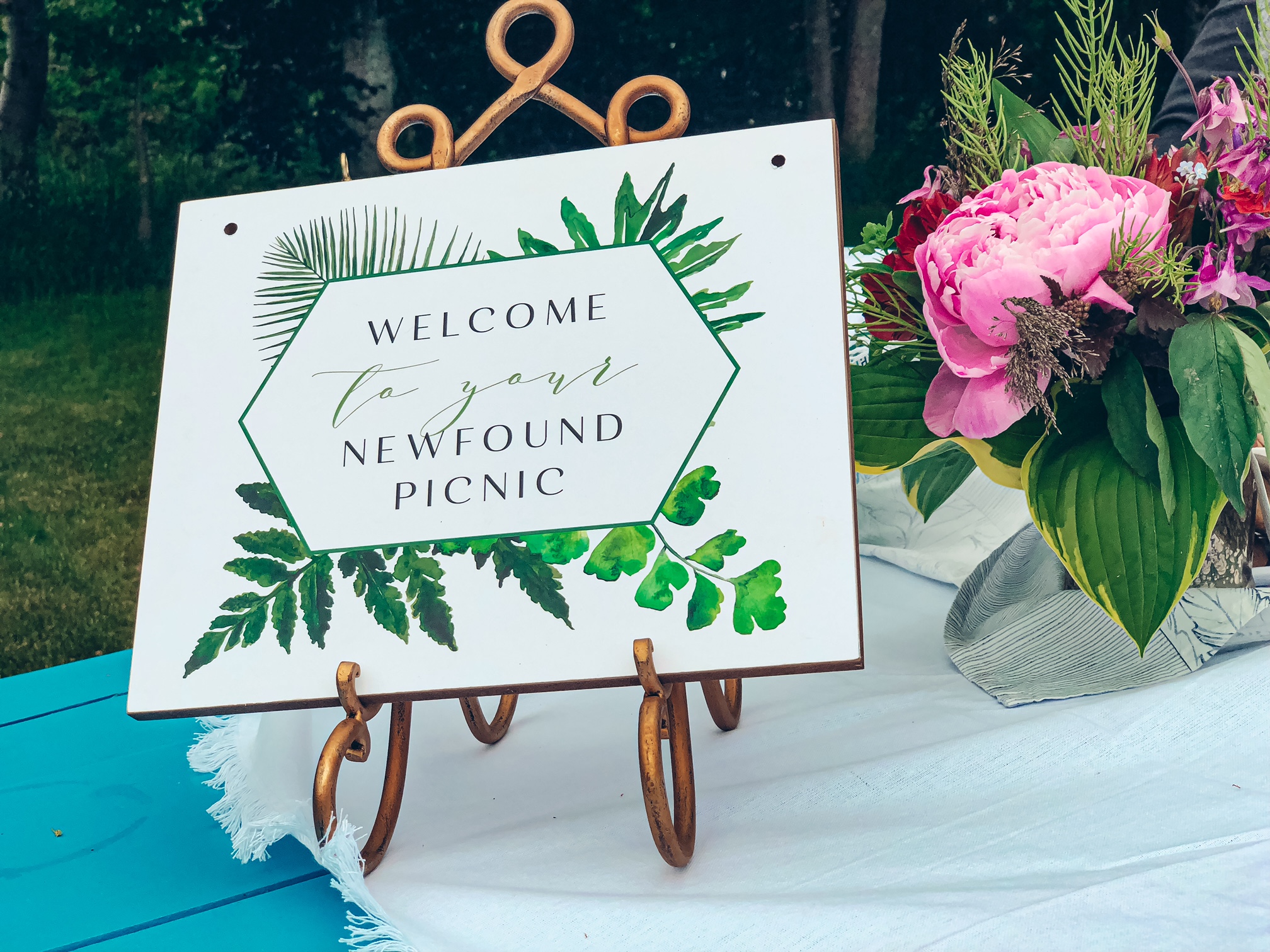 A close up of a white sign that says "Welcome to your Newfound Picnic" with greenery around the text. It sits in a gold sign holder on top of a white tablecloth and there's a bouquet of flowers beside it. 