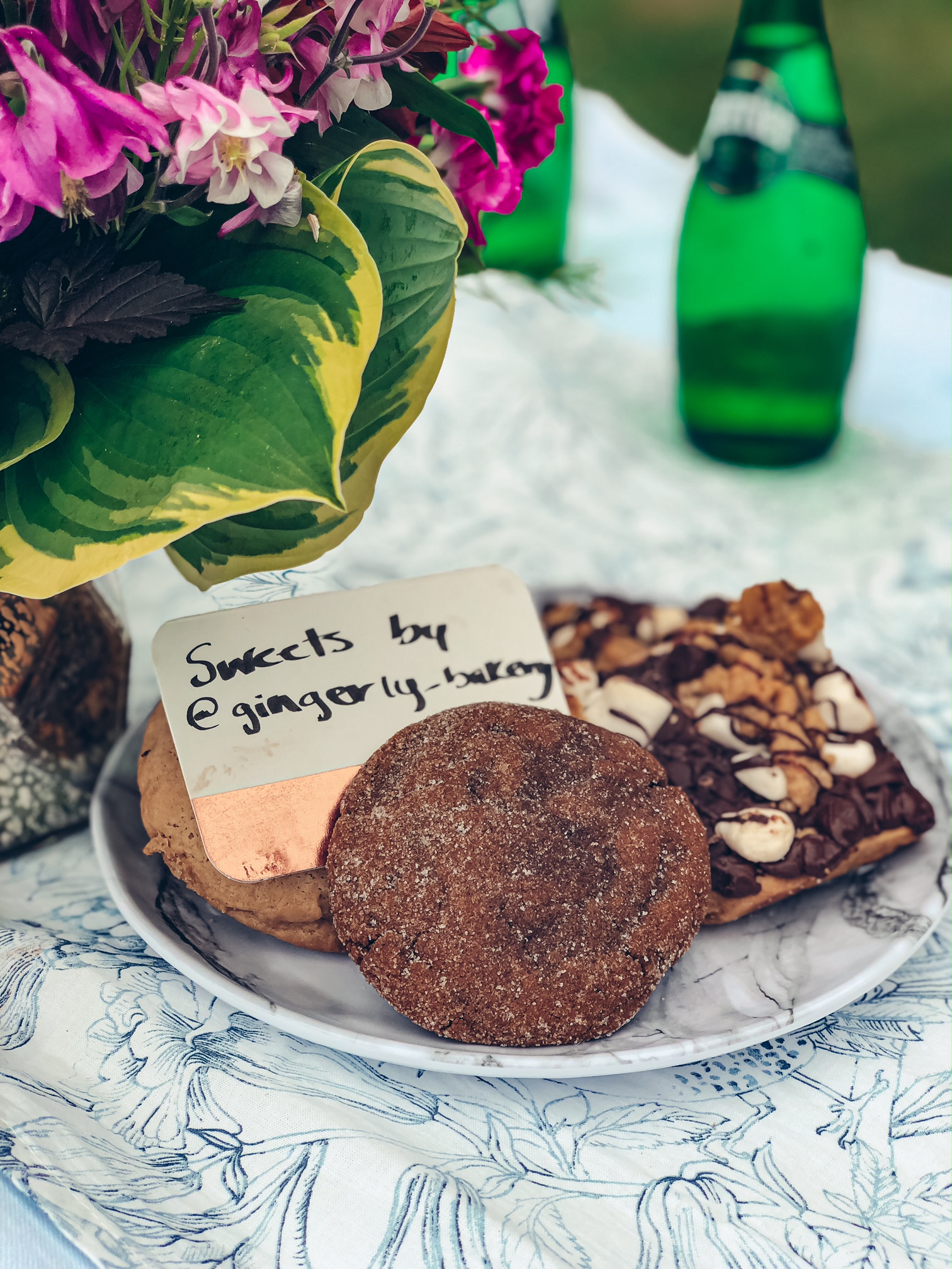 A close up of a plate of Gingerly cookies on top of a white and blue table runner with a bouquet of flowers to the side. 