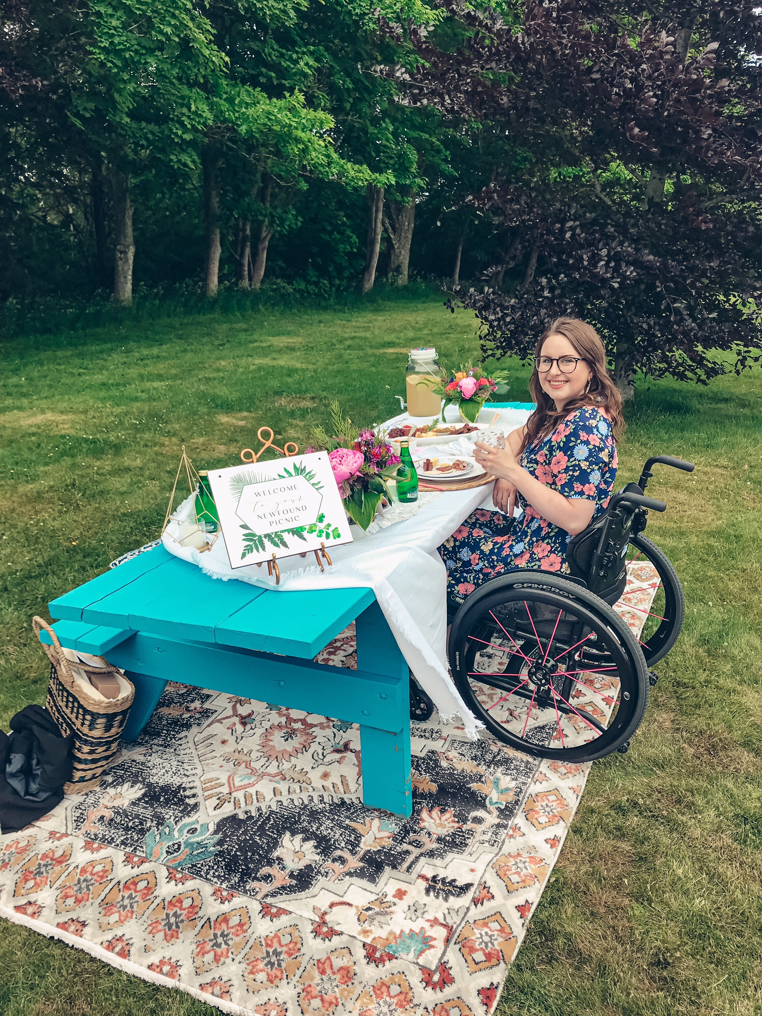 A photo of me sitting in my wheelchair on the accessible side of the picnic table. I'm wearing a navy dress with pink and blue flowers, I'm holding a glass of lemonade and smiling up at the camera.