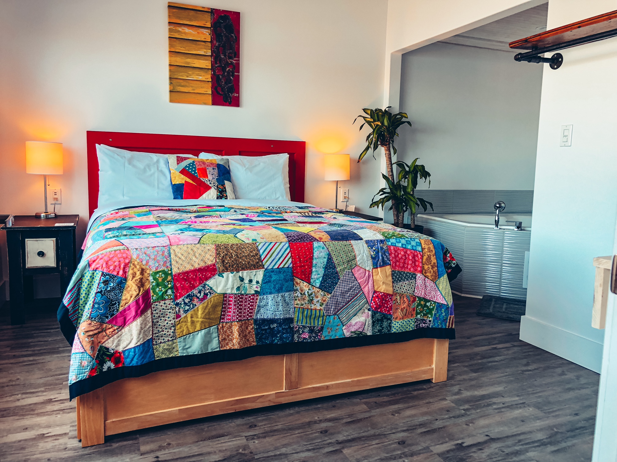A photo of the main bedroom of the suite. There's a queen sized bed with a red headboard and a colourful quilt on it, and in the corner you can see a jacuzzi.
