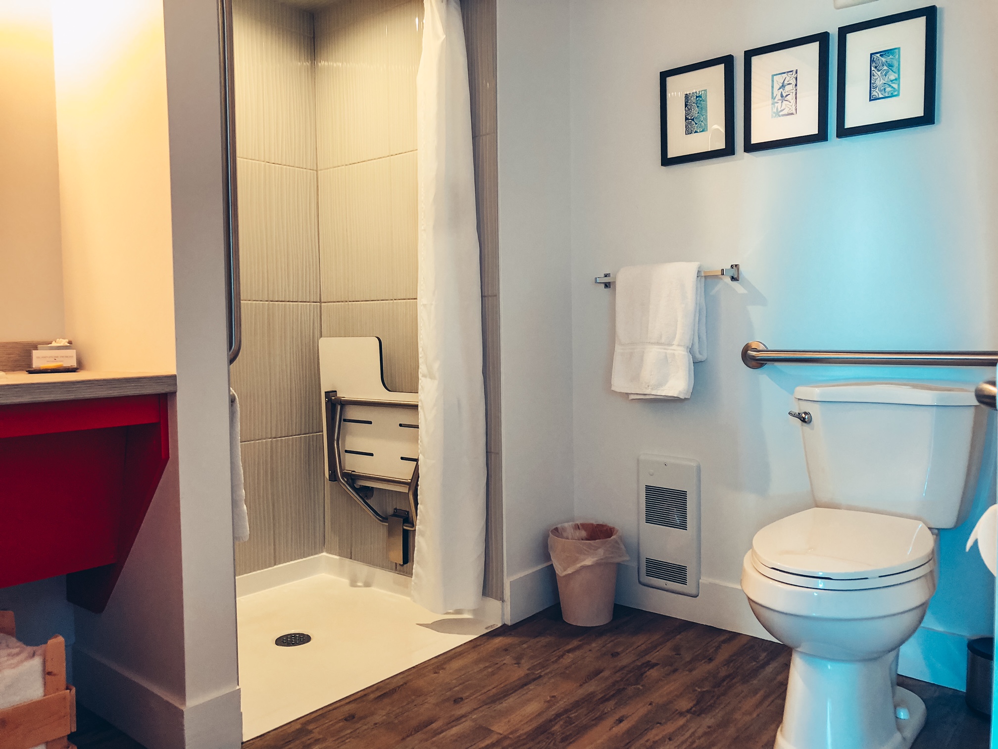 A photo of the bathroom. In the right corner there is a toilet with one of the 2 grabs bars visible. Next to it is a towel rack and beside that is a roll in shower with a bench that is folded up into the wall.