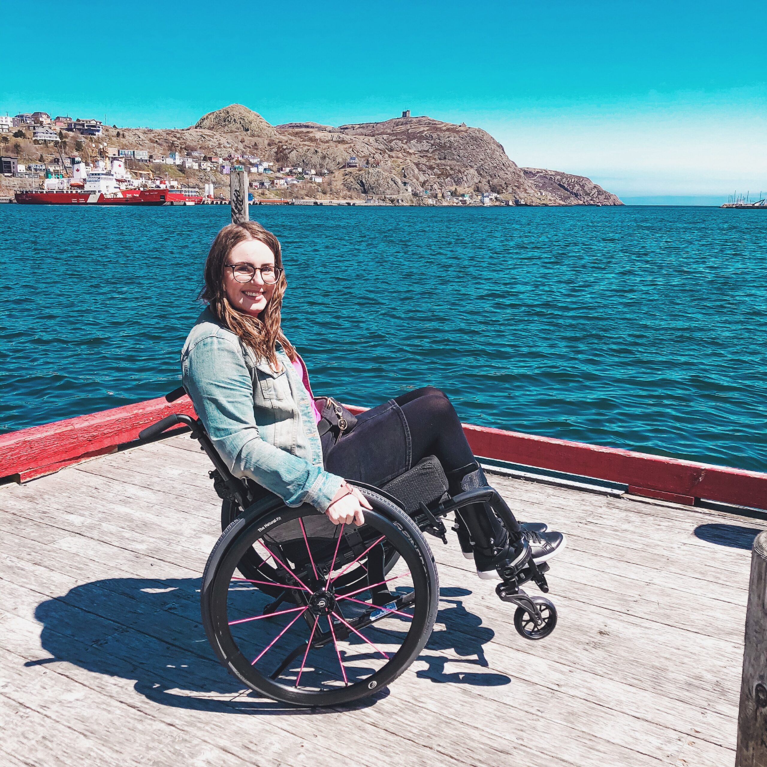 Lisa is doing a wheelie in her wheelchair on a wooden dock at Harbourside Park. In the background is the St. John's harbour and Signal Hill.