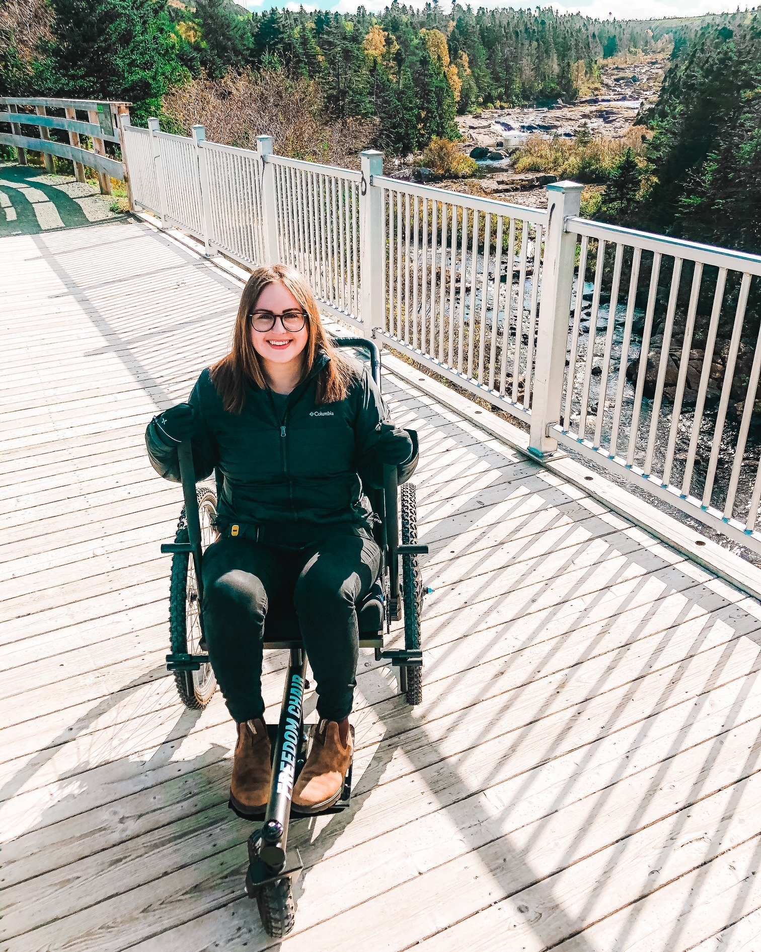A photo of Lisa sitting in the GRIT all terrain wheelchair on a bridge overlooking a river at the Manuel's River Interpretation Centre trail. 