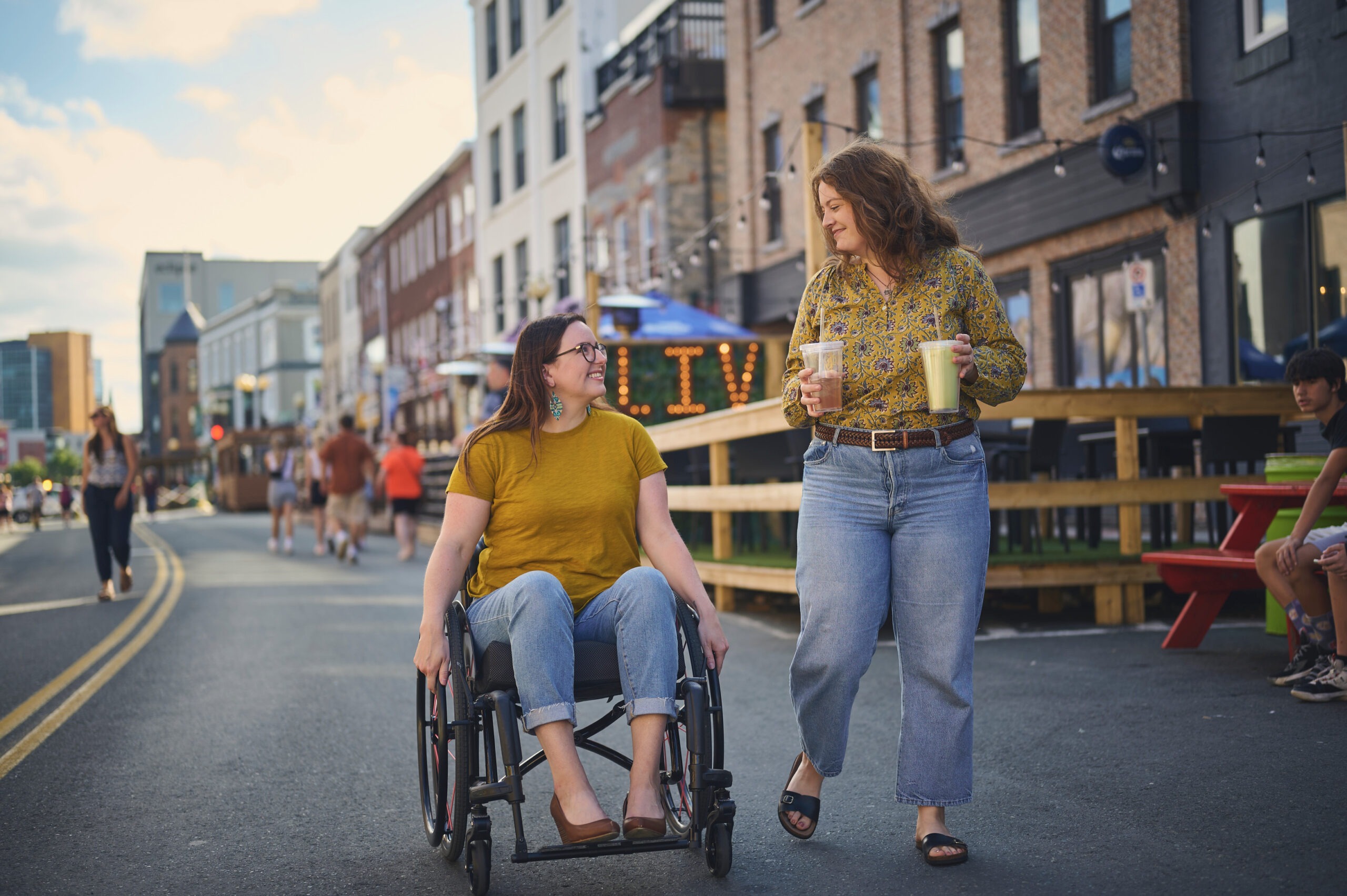 Lisa and her friend Pamela stroll through the pedestrian mall on Water Street. Lisa is in her wheelchair while Pamela walks beside her holding two drinks. 