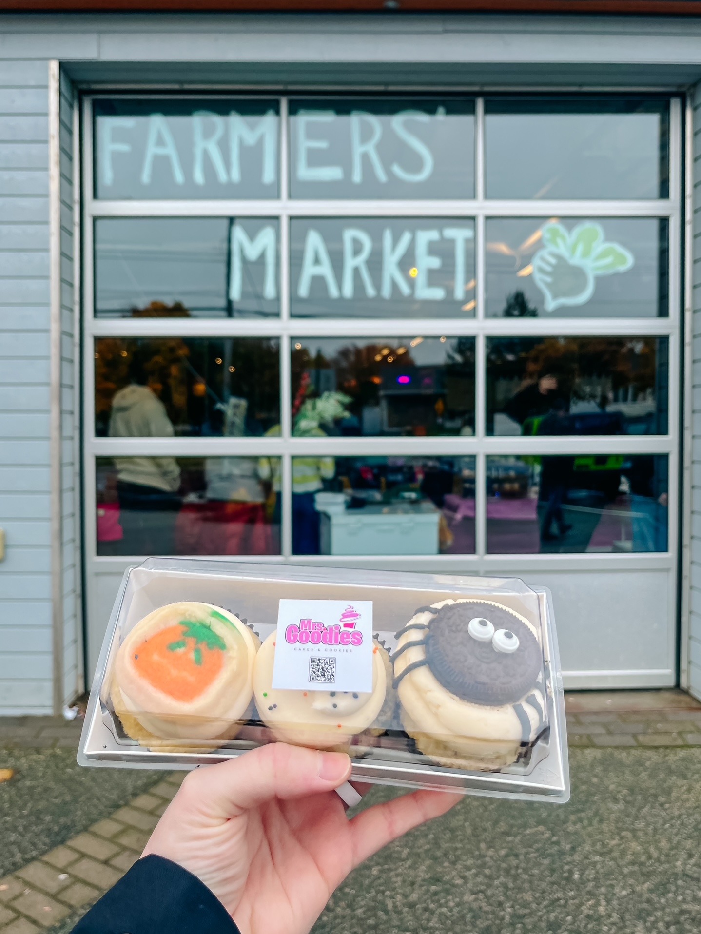 A hand holding a box of three cupcakes in front of the St. John's Farmer's Market.