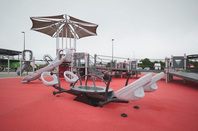 Image of an inclusive playground with red spongey ground, inclusive merry-go-round and a ramped play area in the background. 