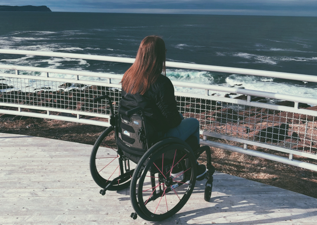 A photo of me from behind, sitting in my wheelchair on a different observation deck near the end of the trail. There are rails in front of me and then cliffs and ocean beyond.