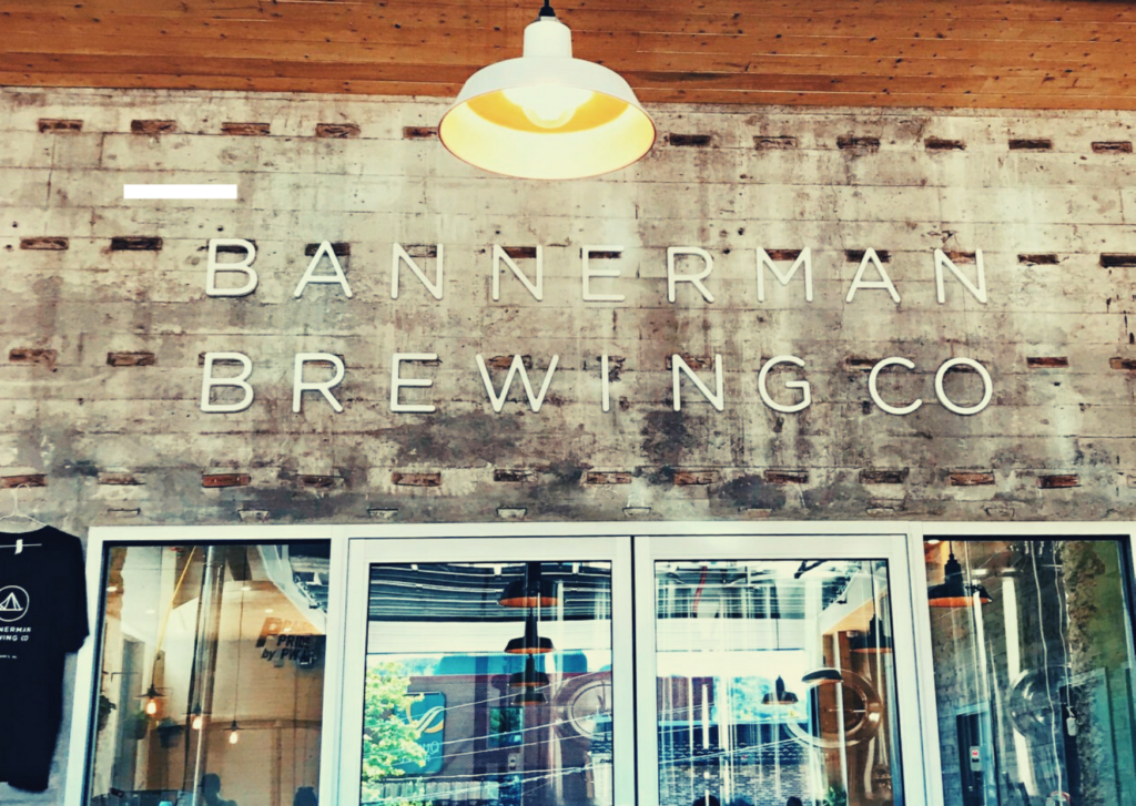 A photo of the back wall of Bannerman Brewing Co. At the top of the wood wall is a sign with the business name. Underneath the sign are glass doors, and through the glass there is brewing equipment visible.